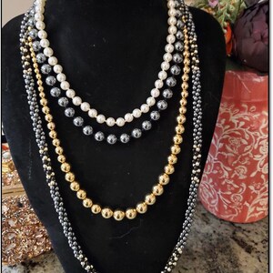 May include: A layered necklace with four strands of beads. The top strand is white pearls, the second strand is dark gray beads, the third strand is gold beads, and the bottom strand is a mix of dark gray and gold beads.