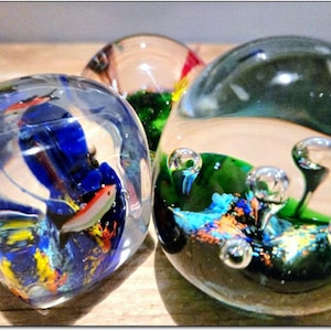 Puede incluir: Tres pisapapeles de vidrio con diseños coloridos. Uno presenta un diseño abstracto azul y amarillo con un pequeño pez. Otro tiene un diseño verde y azul con burbujas. El tercero tiene un diseño rojo, verde y amarillo.