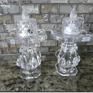May include: Two clear, decorative candle holders with LED candles. Each holder has a faceted base, a decorative midsection, and a hexagonal tray supporting a clear, square candle holder. The LED candles have a flickering flame effect.