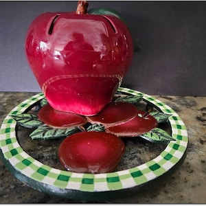 Puede incluir: Una manzana de cerámica roja se sienta sobre un salvamanteles a cuadros verdes y blancos con tres manzanas rojas más pequeñas. El salvamanteles está hecho de hierro fundido.