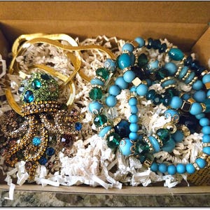 Może przedstawiać: Kartonowe pudełko wypełnione biżuterią, w tym złotym łańcuchem, broszką w kształcie ośmiornicy z klejnotami oraz kilkoma bransoletkami z niebieskich i zielonych koralików. Biżuteria spoczywa na białym wypełniaczu papierowym.