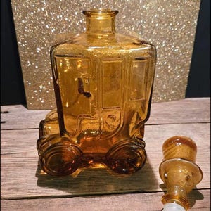Pode incluir: Garrafa de licor de vidro âmbar em forma de carro antigo com um rolha de vidro âmbar separada. A garrafa tem um corpo quadrado e rodas arredondadas.