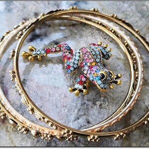 Könnte beinhalten: Ein goldfarbenes Armreif-Set mit einem bunten, mit Juwelen besetzten Frosch-Anhänger. Der Frosch ist mit verschiedenfarbigen Strasssteinen verziert. Die Armreifen haben dekorative Details und liegen auf einer strukturierten Oberfläche.