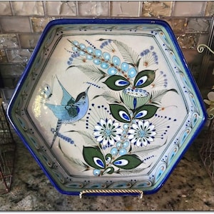 Może przedstawiać: Sześciokątny ceramiczny talerz do serwowania z niebieską obwódką i ręcznie malowanym wzorem kwiatowym i ptasim. Grafika przedstawia niebieskie, zielone i białe kwiaty, kolibra i motyla. Talerz jest podparty złotym metalowym stojakiem.