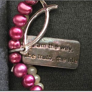 May include: A close-up of a pearl bracelet with a silver clasp and a rectangular charm. The charm is engraved with the words "I am the way, the truth, the life". The pearls are a deep pink color.