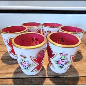 May include: Six white and red glasses with gold rims are arranged on a wooden surface. Each glass features a red interior, floral designs, and red decorative accents. The glasses are likely for serving beverages.