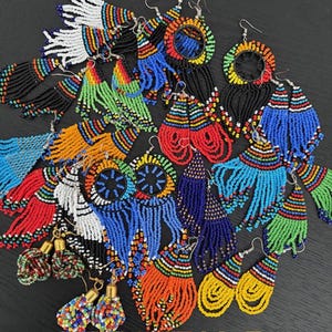 May include: A selection of vibrant beaded earrings in diverse styles. The earrings showcase a spectrum of colours, including red, blue, green, orange, and black. Some have fringing, while others are circular or teardrop-shaped. The earrings are presented on a dark background.
