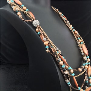 Puede incluir: Un collar de múltiples hilos con una mezcla de cuentas de varios colores y formas. El collar incluye cuentas naranjas, turquesas y marrones, junto con un adorno central brillante. El collar se exhibe en un maniquí negro.