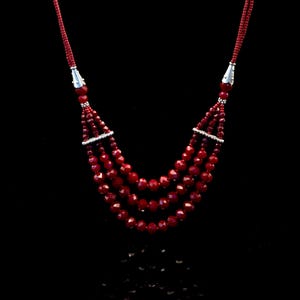 May include: A multi-strand necklace with deep red, faceted beads. The necklace has four layers of beads, connected by silver-coloured accents. The necklace is set against a black background, emphasising the rich colour of the beads.
