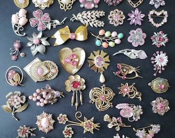 May include: A collection of vintage pink and gold tone brooches featuring various designs including flowers, hearts, bows, and abstract shapes. The brooches are adorned with rhinestones and pearls.