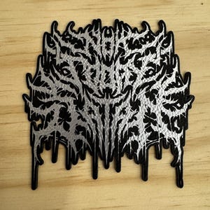 May include: A black and silver patch with a detailed, abstract design. The patch has a black border and a silver, textured interior with intricate patterns. The bottom edge has a dripping effect, adding a unique visual element.
