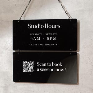 May include: Black rectangular sign with the text "Studio Hours" and business hours. The sign also includes a QR code and the text "Scan to book a session now!" The sign is hanging from a chain.