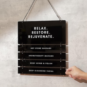 May include: Black acrylic spa menu sign hanging on a gray wall. The sign has the words "RELAX. RESTORE. REJUVENATE." at the top, followed by service options: Hot Stone Massage, Aromatherapy Massage, Body Scrub & Polish, and Deep Cleansing Facial.