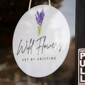 May include: A white round sign with a lavender flower illustration and the text "Wild Flowers Art by Kristine" hanging on a glass door.