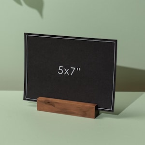 May include: A black 5x7 inch card with white border, displayed in a wooden holder. The card is centered, and the text "5x7" is visible. The holder is a rectangular block of wood.