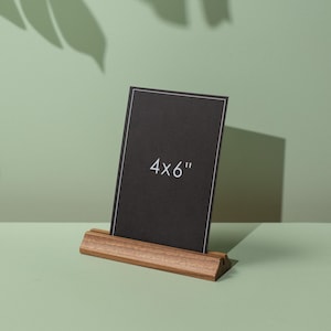 May include: A black card with the text "4x6" is displayed in a wooden holder. The card has a white border. The holder is made of light brown wood. The background is a light green colour.