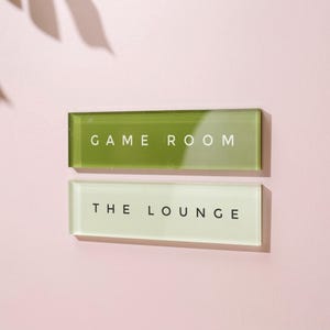 May include: Two rectangular glass signs with white text on a pink wall. The top sign is green and reads "GAME ROOM." The bottom sign is off-white and reads "THE LOUNGE."