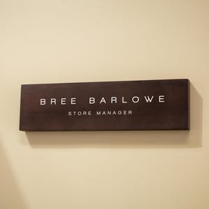 May include: A rectangular wooden sign with the name "BREE BARLOWE" and the title "STORE MANAGER" in white lettering. The sign is a dark brown color and is mounted on a light beige wall.