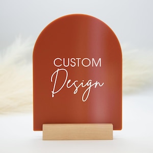 May include: A terracotta-colored arched sign with the words "CUSTOM Design" in white script. The sign is mounted on a small, rectangular wooden base, suitable for display. The background is a soft, neutral color.