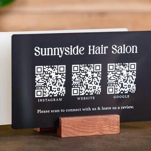 May include: Black sign with white text that reads "Sunnyside Hair Salon". The sign has three QR codes labeled "Instagram", "Website", and "Google". The sign is on a wooden stand and has the text "Please scan to connect with us & leave us a review."