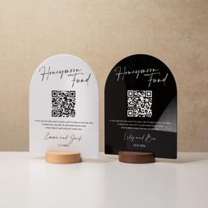 May include: Two arched signs with wooden bases. One is white with black text that reads "Honeymoon Fund" and includes a QR code. The other is black with white text. Both signs have a message and names.