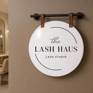 May include: A white, circular sign for "The Lash Haus Lash Studio" hangs on a brown wall. The sign is suspended by brown leather straps from a black metal rod. The interior of a salon is visible in the background.