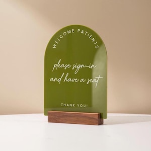 May include: A green arched sign with the words "WELCOME PATIENTS" at the top and "please sign-in and have a seat" in the center. The sign is mounted on a rectangular wooden base. The words "THANK YOU!" are at the bottom.
