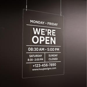 May include: A clear, rectangular sign with the words "WE'RE OPEN" in bold white letters. The sign also displays business hours, a phone number, and a website address. The sign is suspended by thin wires against a dark background.