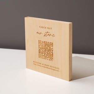 May include: A light-colored wooden block with engraved text and a QR code. The text reads "CHECK OUT our store" and "MODERN ALBUM DESIGNS" with a website address. The block is on a white surface with a gray background.
