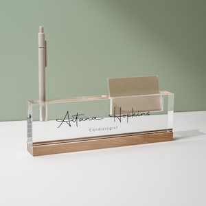 May include: Clear acrylic desk organizer with a wooden base. It holds a pen, business cards, and features the name "Aitana Hopkins, Cardiologist" in black script. The background is a soft green.