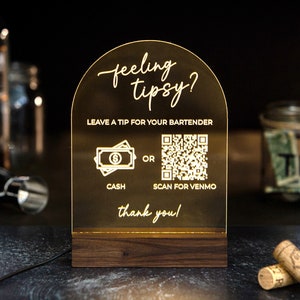 May include: A gold-colored acrylic sign with a rounded top that reads "feeling tipsy?" and has a QR code and a cash icon with the text "Leave a tip for your bartender, OR, Cash, Scan for Venmo, thank you!"
