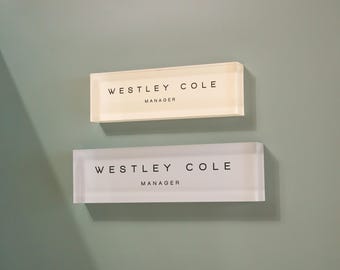 May include: Two rectangular nameplates are mounted on a wall. The top one is off-white, the bottom one is clear. Both read "WESTLEY COLE MANAGER" in black sans-serif font. The wall is a muted green color.