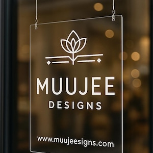 May include: Clear acrylic sign with the white text "MUUJEE DESIGNS" and a logo of a stylized flower. The website address www.muujeedesigns.com is also displayed. The sign is suspended by thin metal wires.