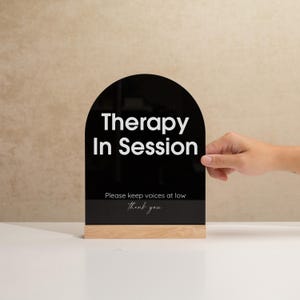 May include: A black acrylic sign with the words "Therapy In Session" in white, atop a light wood base. The sign also includes the text "Please keep voices at low" and "thank you".