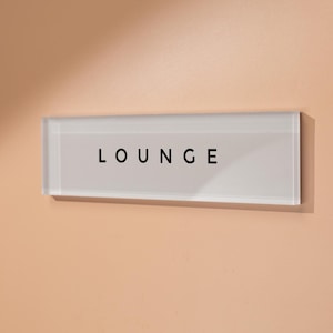 May include: A rectangular white sign with the word "LOUNGE" in black letters is mounted on a peach-colored wall. The sign is made of a clear, glass-like material. The wall is illuminated by natural light.