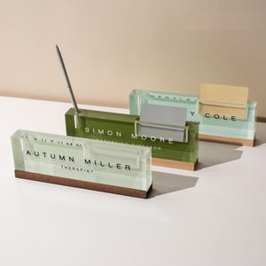 May include: Three rectangular desk name plates made of clear acrylic with wooden bases. Each features a name and title printed in black text, including Autumn Miller, Simon Moore, and Wesley Cole, with a silver pen in one holder.