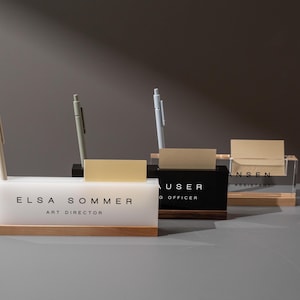 May include: A collection of desk organizers with pens and business card holders. The organizers are made of acrylic, wood, and black materials. The text on the organizers reads "ELSA SOMMER ART DIRECTOR", "HAUSER G OFFICER", and "HANSEN ASSISTANT".