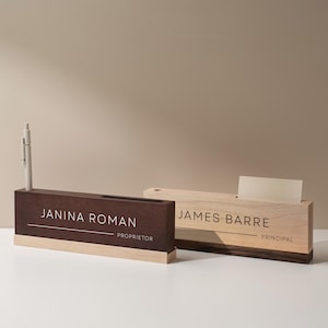 May include: Two wooden desk organizers. One is dark brown with a light wood base, holding a pen and labeled "Janina Roman, Proprietor." The other is light wood, labeled "James Barre, Principal."