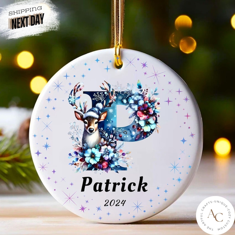 May include: A round white ceramic ornament featuring a large blue letter P decorated with blue and purple flowers and a deer wearing a winter hat. The name Patrick and the year 2024 are printed in black below the letter, surrounded by small blue stars.
