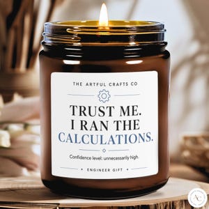 May include: A brown glass jar candle with a lit wick and a white label. The label reads: THE ARTFUL CRAFTS CO, TRUST ME. I RAN THE CALCULATIONS., Confidence level: unnecessarily high., ENGINEER GIFT. The text is in black and blue ink.