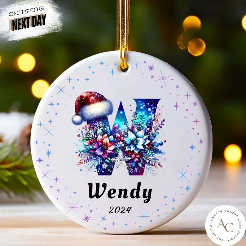 May include: A round white ceramic ornament featuring a large letter W decorated with colourful floral patterns and a Santa hat. The name Wendy and the year 2024 are printed below the letter. The ornament is accented with blue and purple star motifs.
