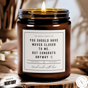 May include: A lit brown glass jar candle with a gold rim and a white label. The label reads "YOU SHOULD HAVE MOVED CLOSER TO ME, BUT CONGRATS ANYWAY :)". The candle is labeled as vegan and 100% organic.