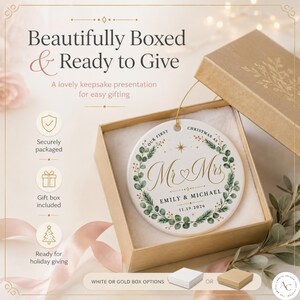 May include: A round white ceramic ornament with a gold string, featuring a green eucalyptus wreath design, the text "Our First Christmas As Mr & Mrs", "Emily & Michael", and "11.19.2026". It is presented in a gold gift box with a lid.