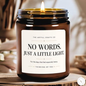May include: A brown glass jar candle with a lit wick. The white label features the text: THE ARTFUL CRAFTS CO, NO WORDS. JUST A LITTLE LIGHT. For the days that feel especially heavy. THINKING OF YOU. The candle emits a warm glow from the flame.