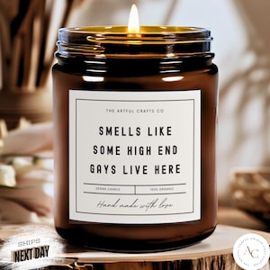 Puede incluir: Un tarro de vela de vidrio marrón con una etiqueta blanca que dice "The Artful Crafts Co. Smells Like Some High End Gays Live Here. Vegan Candle 100% Organic Hand made with love."