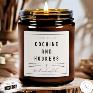 May include: A brown glass jar candle with a lit wick and a white rectangular label. The label features the text THE ARTFUL CRAFTS CO, COCAINE AND HOOKERS, VEGAN CANDLE, 100% ORGANIC, and Hand made with love in black sans-serif and script fonts.