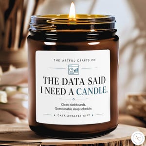 May include: A brown glass jar candle with a lit wick. The white label features the text: THE DATA SAID I NEED A CANDLE. Clean dashboards. Questionable sleep schedule. DATA ANALYST GIFT. The Artful Crafts Co logo is at the top with a chart icon.