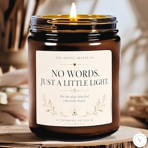 May include: A lit candle in a dark amber glass jar with a white label featuring the text "THE ARTFUL CRAFTS CO", "NO WORDS. JUST A LITTLE LIGHT.", "For the days that feel especially heavy.", and "THINKING OF YOU" with decorative botanical line art.