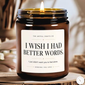 May include: A brown glass jar candle with a lit wick and a white rectangular label. The label features the text: THE ARTFUL CRAFTS CO, I WISH I HAD BETTER WORDS., I just didn't want you to feel alone., and SENDING YOU LOVE, with small diamond accents.
