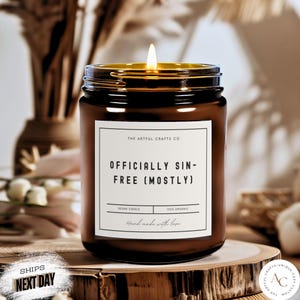 Könnte beinhalten: Ein braunes Glas-Kerzengefäß mit einem weißen Etikett, auf dem "The Artful Crafts Co. Officially Sin-Free (Mostly)" steht. Das Etikett trägt außerdem die Aufschrift "Vegan Candle" und "100% Organic". Die Kerze ist angezündet und das Gefäß steht auf einer Holzoberfläche.